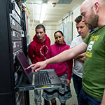 four people standing in from of laptop screen. One of the people pointing at screen.