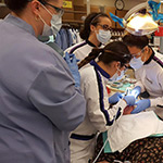 Five people in a dental clinic. One of the people is laying down in a dental chair with a towel on their chest. Two of the people are wearing masks over their mouths and noses and working on the person in the dental chair with tools. The other two people are standing near the two people working on the person in the dental chair and observing. There is screen near them showing part of the inside of the mouth of the person being worked on.