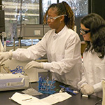 Two people in lab coats with protective goggles on in a lab working with lab equipment.